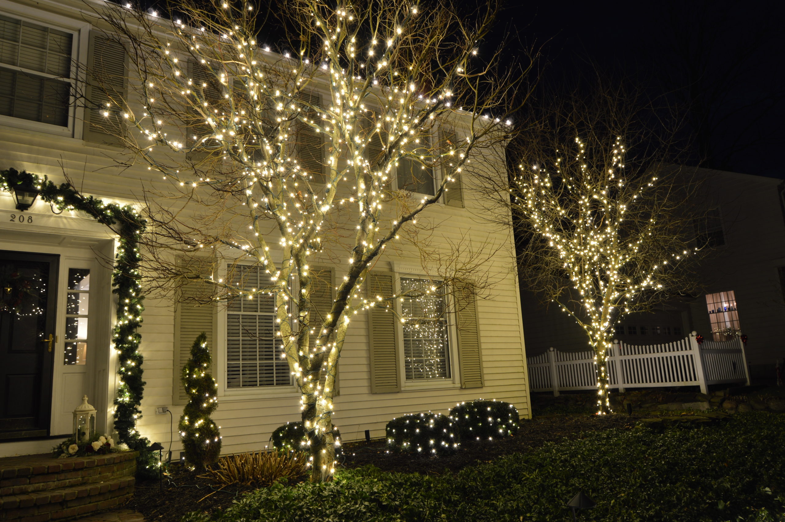 A white house with holiday lighting on trees and shrubbery